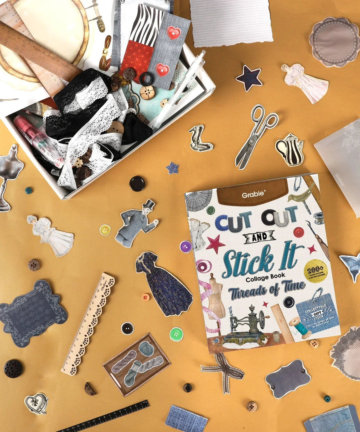 Art supplies and scrapbook cutouts surrounding the Threads Of Time-Themed Grabie Scrapbook Club Box on tan background