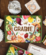 The Flavor Folio-Themed Grabie Scrapbook Club Box with colorful food art and art supplies on wooden table