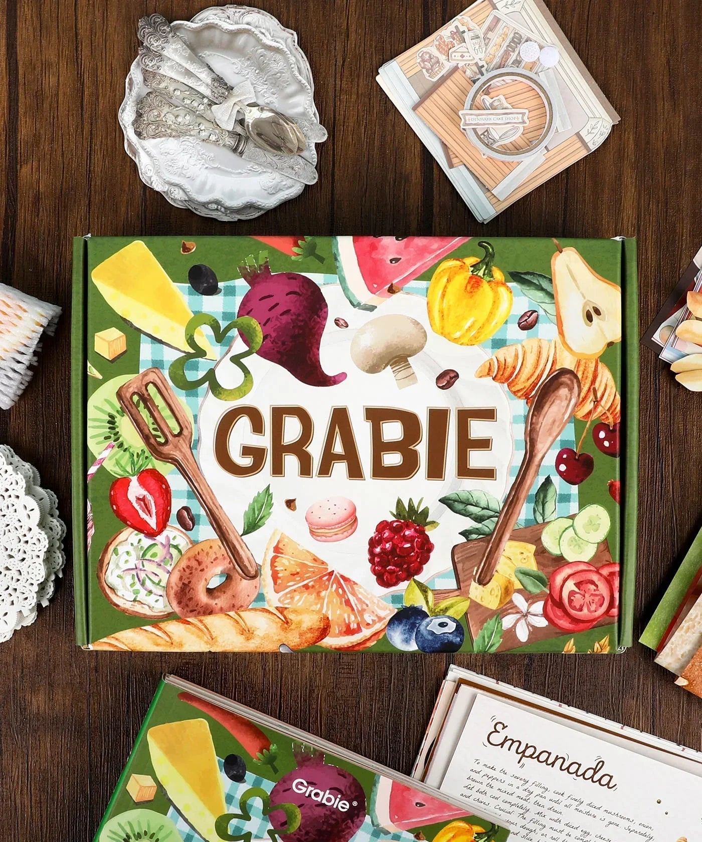 The Flavor Folio-Themed Grabie Scrapbook Club Box with colorful food art and art supplies on wooden table