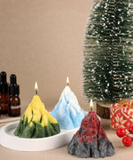 Three lit mountain-shaped candles in yellow-green, blue, and red-black on a tray with a festive tree and gift behind, Scent Candle DIY Set