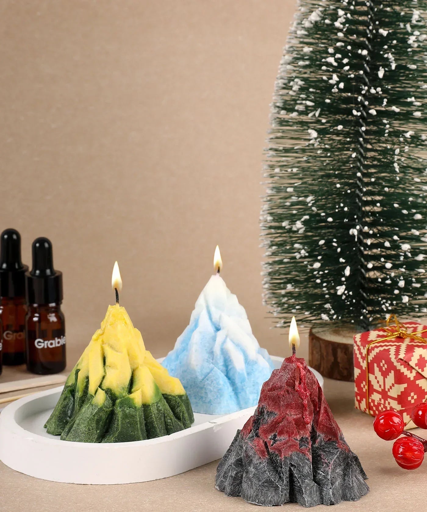 Three lit mountain-shaped candles in yellow-green, blue, and red-black on a tray with a festive tree and gift behind, Scent Candle DIY Set