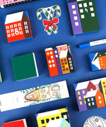 Colorful boxes with winter and city designs arranged with a pen and candy cane for the 2025 Limited Edition Holiday Advent Calendar