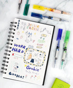 Dual-Tip Notetaking Pen Set Of 6 with colorful drawings and lettering on a sketchbook on white marble background