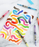 Dual-Tip Notetaking Pen Set Of 6 with colorful worms drawn on paper surrounded by translucent pens on marble surface