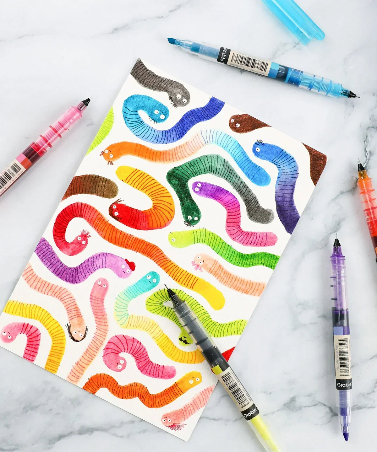 Dual-Tip Notetaking Pen Set Of 6 with colorful worms drawn on paper surrounded by translucent pens on marble surface