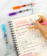 Hand using pink pen from Dual-Tip Notetaking Pen Set Of 6 to write colorful notes in a spiral notebook with art supplies nearby