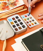 Watercolor Pocket Set of 12 - Autumn Colors with brushes, a color chart, and art supplies on an orange background
