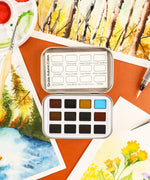 Watercolor Pocket Set of 12 - Autumn Colors open with vibrant paint pans on a desk surrounded by autumn-themed watercolor paintings and brushes