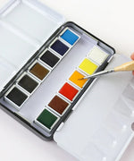 Grabie x @SSavaArt Watercolor 12 Half Pan Deluxe Travel Set with brush dipping into vibrant orange paint for artists.