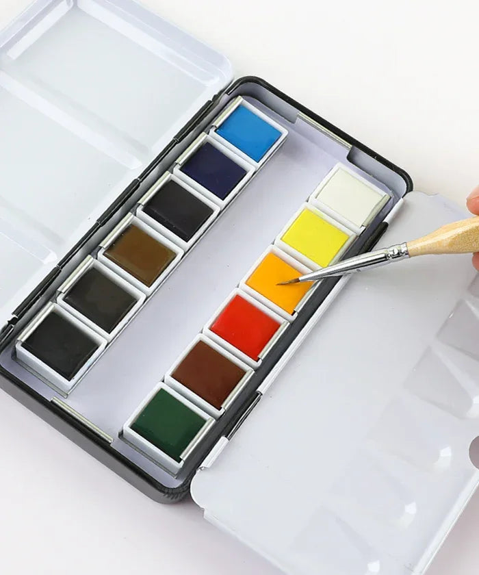 Grabie x @SSavaArt Watercolor 12 Half Pan Deluxe Travel Set with brush dipping into vibrant orange paint for artists.