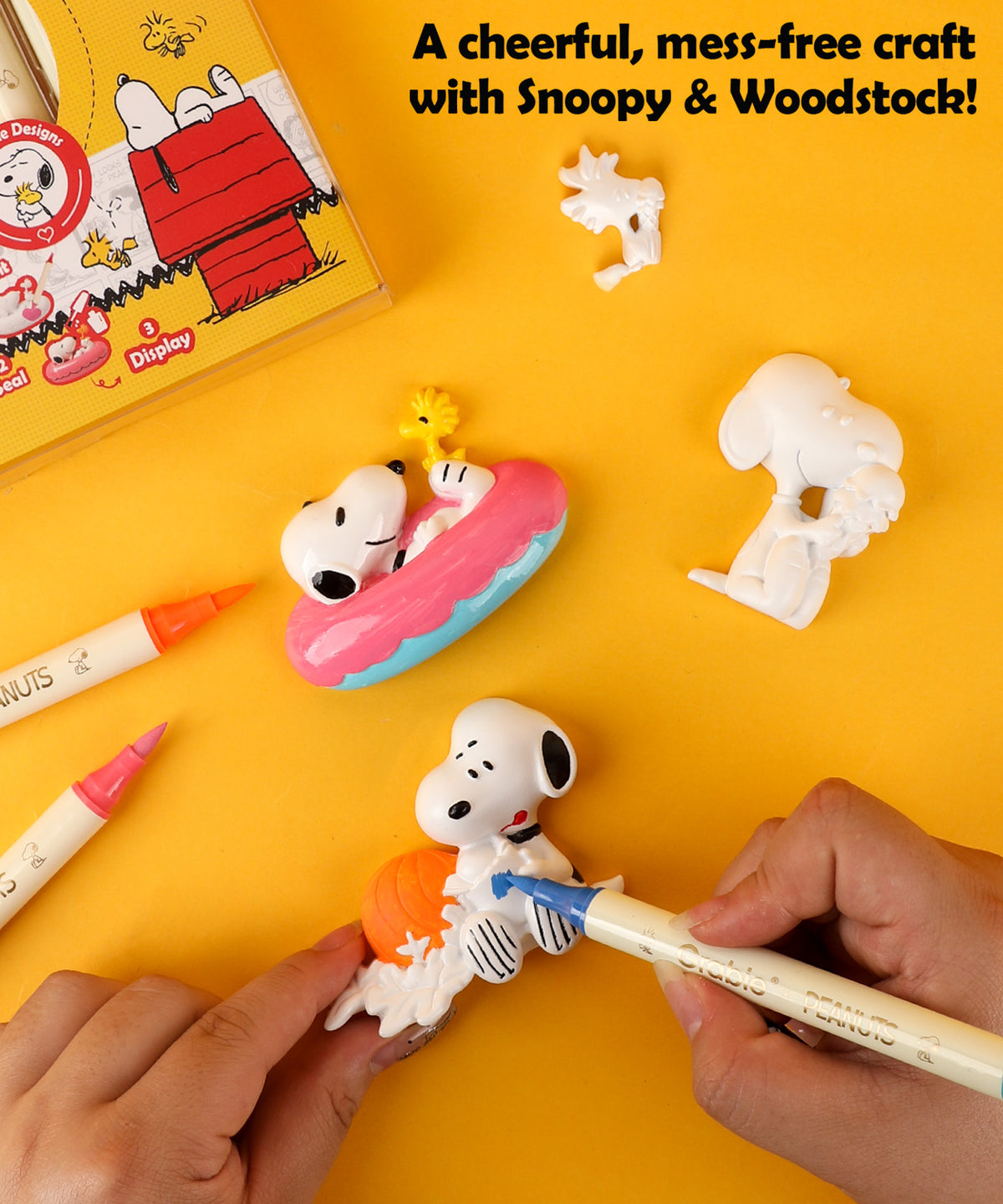 Hands painting a Snoopy and Woodstock resin figure with blue and orange dual tip markers.