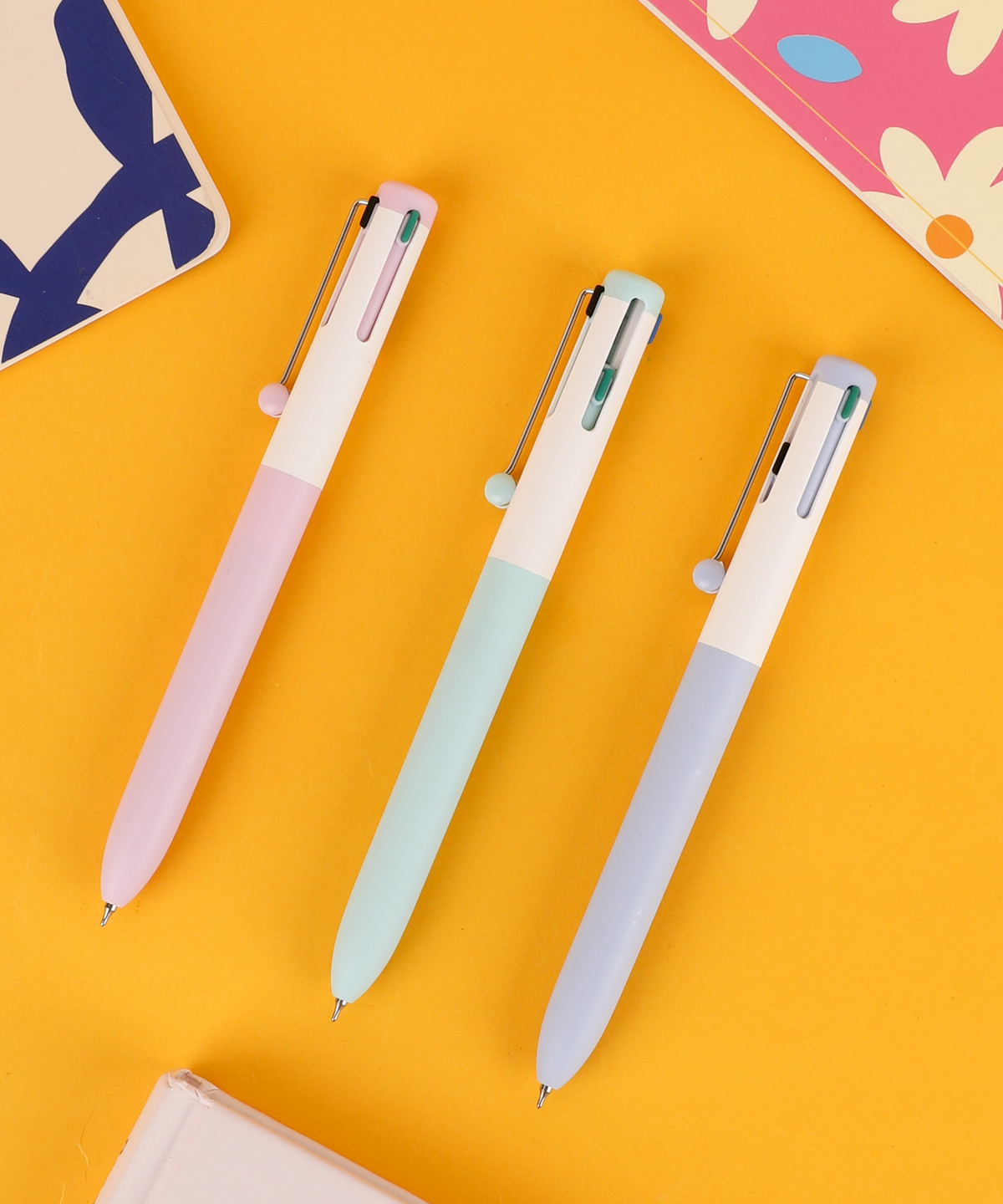 Set of 3 pastel 4 color pens in pink, light blue, and lavender lying neatly on a bright yellow surface.
