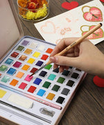 Hand holding 11 Pcs Miniature Detail Paint Brush Set With Natural Wood Handle dipping into a colorful watercolor palette next to art sketches.